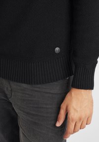 Maglione nero lavorato a maglia con un motivo testurizzato, polsini e orlo a costine, con un piccolo dettaglio del logo argento nella parte inferiore.