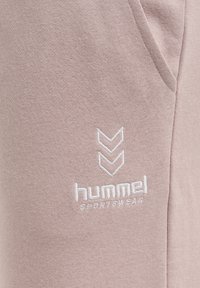 Hellrosa Stoff mit einer Seitentasche und weiß besticktem "hummel SPORTSWEAR"-Logo mit zwei nach unten zeigenden Chevronen.