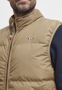 Beige puffertaki, jossa on korkea kaulus ja vetskari, jossa on tummansinisiä hiusosia ja pieni kolmivärinen logo rinnassa.