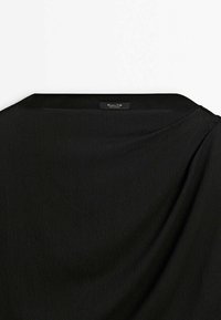 Vêtement noir avec col bateau et texture visible du tissu, montrant une petite étiquette de marque rectangulaire cousue à l'intérieur du col arrière.
