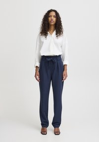 Chemise blanche à boutons, associée à un pantalon taillé marine à taille nouée, avec une texture lisse et des jambes larges. Le mannequin se tient droit, portant des talons minimalistes.