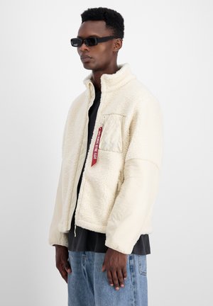 Cremfarbener Blazer mit einem flauschigen, texturierten Außenmaterial, glatten Ärmeln, einer Utility-Tasche und einem roten Etikett. Trägt man über einem schwarzen Shirt.