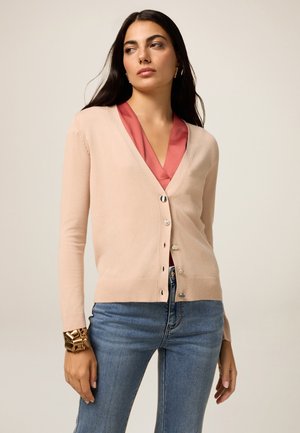 Donna che indossa un cardigan beige chiaro abbottonato sopra una blusa rosa, abbinata a jeans blu e a un grande bracciale d'oro sul polso sinistro.