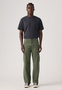 Ανδρικό χακί cargo παντελόνι με πλαϊνές τσέπες, συνδυασμένο με σκούρο T-shirt και μαύρα αθλητικά παπούτσια. Το ύφασμα είναι απαλλαγμένο, με σχεδιασμό ίσιας κοπής.