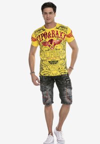 Geel grafisch T-shirt met rode ontwerpen en patronen, gecombineerd met grijze cargoshorts met zakken en rode stiksels. Witte sneakers.