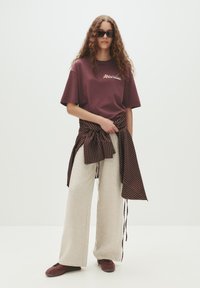 Kastanjebrun t-shirt med logo, beige vide bukser og et kastanjebrun og stribet bælte bundet om taljen. Brune sneakers. Krøllet hår og solbriller.