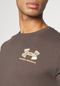 Brun bomulls t-shirt med en kamouflage Under Armour-logotyp i gult och vitt. Kort ärm och design med rund hals.