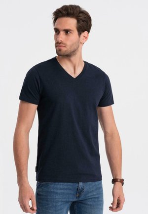 T-shirt bleu marine à col en V fabriqué en tissu doux, avec des manches courtes et une coupe ajustée. Porté avec un jean bleu et une montre marron au poignet.