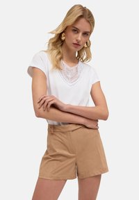 Camiseta blanca de manga corta con un escote decorativo de encaje, combinada con pantalones cortos de talle alto en tono marrón claro, confeccionados con una tela suave.