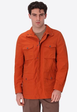 Giovane uomo con capelli ricci scuri che indossa una giacca arancione con quattro tasche frontali con patta sopra una camicia beige, in piedi e rivolto in avanti.