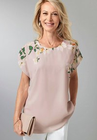 Rosa Bluse mit floralem Design an den Ärmeln, aus leichtem Stoff. Das Model hält eine beige Clutch; der Hintergrund ist neutral grau.