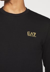 Primer plano de una persona que lleva una camiseta negra con un pequeño logo amarillo de "EA7 Emporio Armani" en el lado izquierdo del pecho.