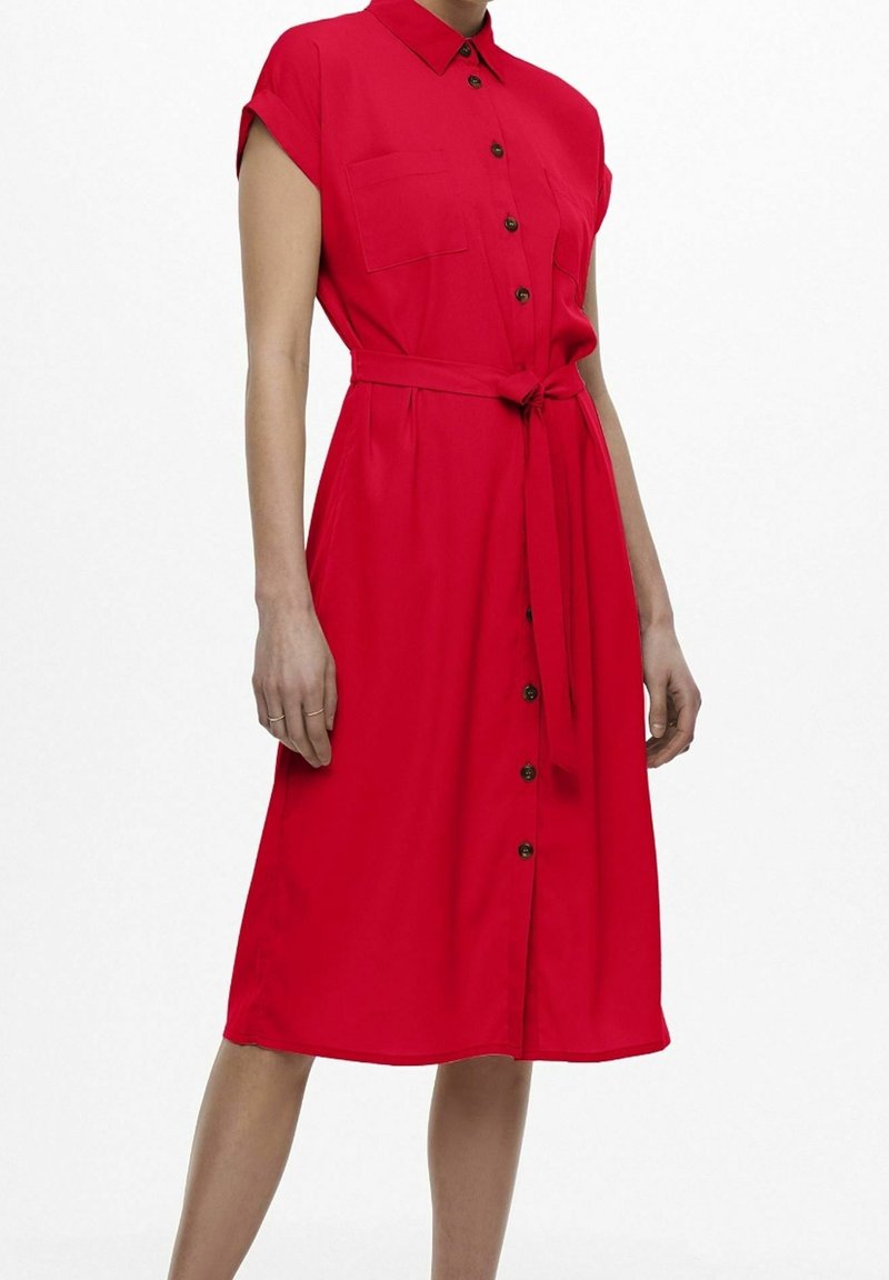 Femme portant une robe rouge boutonnée jusqu'aux genoux, à manches courtes, avec des poches sur la poitrine et une ceinture nouée à la taille.