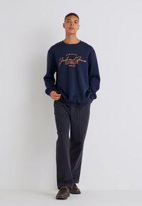 Navy sweatshirt with orange logo print, paired with dark pinstriped trousers. Model is wearing brown shoes. Minimalistic, casual outfit.