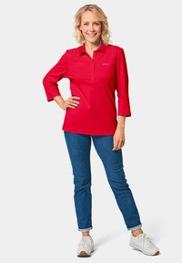 Rotes Polohemd mit Kragen und Dreiviertelärmeln, kombiniert mit schmalen blauen Jeans und weißen Sneakers. Einfaches Design und lässige Passform.
