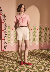 Růžové tričko s potiskem třešní, bílé lněné šortky, hnědý pásek a červené síťované baleríny, vše na pozadí s květinovým vzorem.