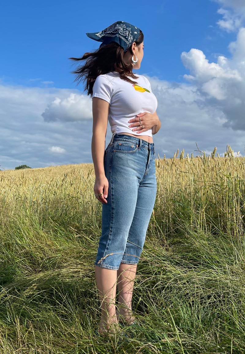 T-shirt blanche à manches courtes avec un graphique de citron, associée à un jean cropped bleu clair, dans un champ naturel avec de l'herbe verte et un ciel bleu.