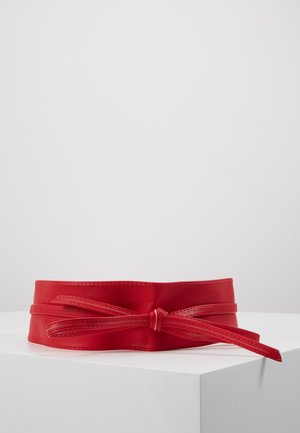 Ceinture large en cuir rouge avec un nœud noué à l'avant, exposée sur un socle blanc sur un fond uni.