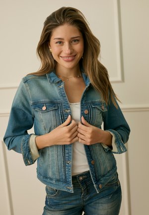 Mladá žena se usmívá, nosí dvoubarevnou džínovou bundu přes bílý top a džíny, ruce drží přední část bundy, stojí uvnitř u krémové stěny.