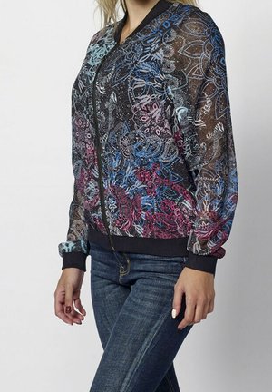 Femme portant une veste bomber noire transparente avec des motifs floraux multicolores et des motifs paisley, associée à un jean bleu en denim.
