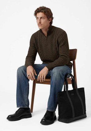 Un homme est assis sur une chaise en bois. Il porte un pull marron à torsades, un jean et des chaussures noires ; un sac cabas noir se trouve à côté de lui.