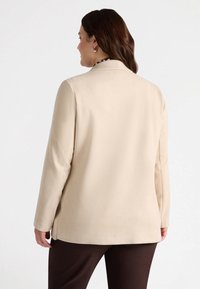 Blazer beige con una texture liscia, maniche lunghe e un colletto a punta; presenta fessure laterali per un comfort aggiuntivo e una vestibilità rilassata.
