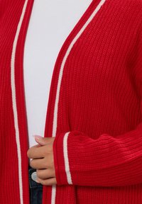 Röd stickad cardigan med vita sidostripes, som har en lös passform och ribbad textur. Inkluderar en veckad mudd och en öppen framdesign.