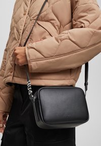 Person iført en beige quiltet jakke og sorte bukser, der holder en sort HUGO læder crossbody taske med mærket remdetalje.