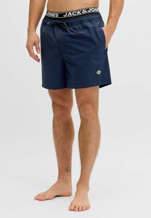 Uomo che indossa pantaloncini da bagno blu navy con fascia elastica in vita e biancheria intima nera e bianca marchiata "Jack & Jones" visibile.