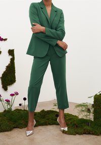 Traje verde a medida hecho de tela suave, con un blazer de un solo botón y pantalones recortados, combinado con tacones puntiagudos plateados.