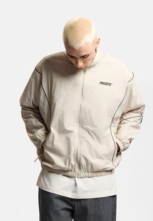 Młoda osoba z krótkimi blond włosami patrzy w dół, ubrana w beżową kurtkę na zamek z logo "PROSTO" i z rękami w kieszeniach.