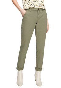 Donna che indossa pantaloni verde oliva arrotolati, una blusa floreale e stivali con tacco bianchi alla caviglia, in piedi con una mano in tasca.