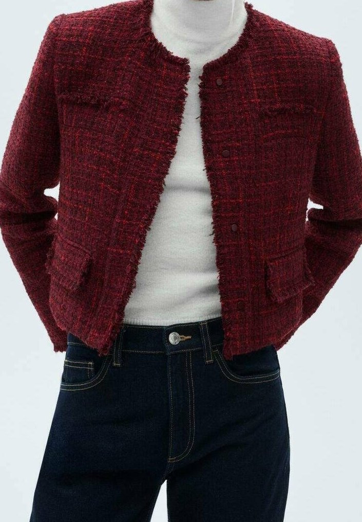 Femme portant une veste courte texturée rouge foncé sur un col roulé blanc et un jean bleu foncé sur un fond clair.