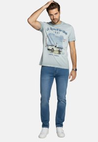 Camiseta azul claro con un estampado gráfico de un coche vintage, combinada con jeans de mezclilla de lavado medio y zapatillas blancas. El ajuste es casual y cómodo.