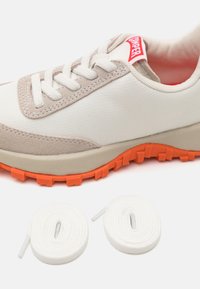 Białe sneakersy z beżowymi zamszowymi akcentami, teksturowanym materiałem, pomarańczową gumową podeszwą i płaskimi białymi sznurowadłami ułożonymi w pętle obok buta.