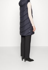 Chaleco acolchado azul marino con capucha, con un patrón de cheurón, cierre frontal con cremallera y un pequeño detalle de logo. Llevado sobre una camiseta de manga larga y pantalones.