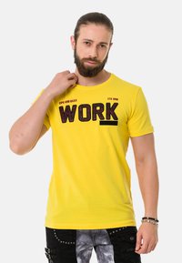 Gelbes Baumwoll-T-Shirt mit dem Wort "WORK" in fettem schwarzen Text, Rundhals, kurzen Ärmeln und taillierter Passform. Das Modell trägt schwarze Hosen.