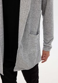Hombre vestido con un cárdigan claro de frente abierto y con bolsillo sobre una camiseta negra y pantalones negros, de pie contra un fondo blanco liso.