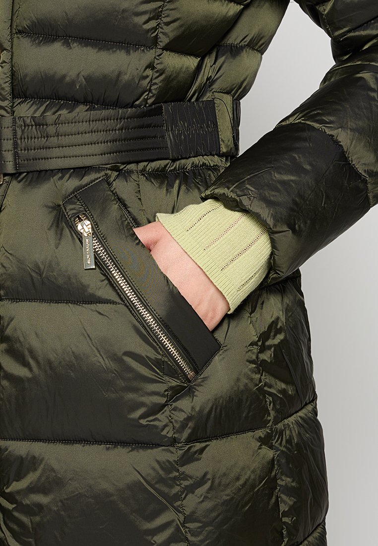 Hand in einer hellgrünen gestrickten Ärmeln in einer mit Reißverschluss versehenen Tasche einer dunkelolivgrünen gesteppte Pufferjacke mit Taillengürtel.