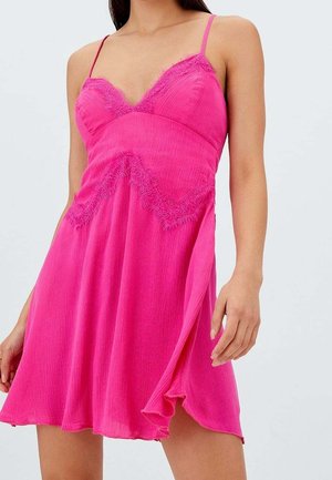 Vrouw draagt een felroze mouwloze jurk met kanten rand en een vloeiende rok tegen een effen lichte achtergrond.