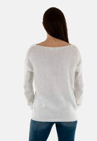 Suéter blanco de punto con diseño de espalda descubierta, mangas largas y un corte relajado, combinado con jeans azules. Textura suave, estilo casual.