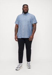 Camisa de botón de manga corta, de color azul claro, en una tela texturizada, combinada con pantalones negros y zapatillas grises. El modelo está de pie de frente.
