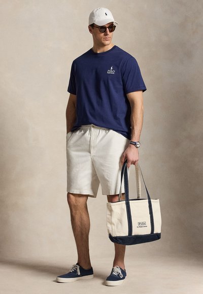 Man wearing navy Polo Ralph Lauren t-shirt, beige shorts, white cap, sunglasses, navy sneakers, holding a beige and navy tote bag.