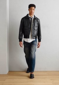 Schwarze Lederbomberjacke mit zwei Fronttaschen, getragen über einem weißen Hemd und einer schwarzen Krawatte. Blue Jeans und schwarze Schuhe runden das Outfit ab.
