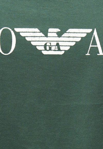 Λευκός στυλιζαρισμένος αετός με απλωμένα φτερά και τα γράμματα "G A" στο κέντρο, σε φόντο πράσινου υφασμάτινου υλικού με υφή.