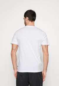 Hombre de espaldas vistiendo una camiseta blanca de manga corta y pantalones cortos negros, con tatuajes visibles en ambos brazos sobre un fondo blanco.