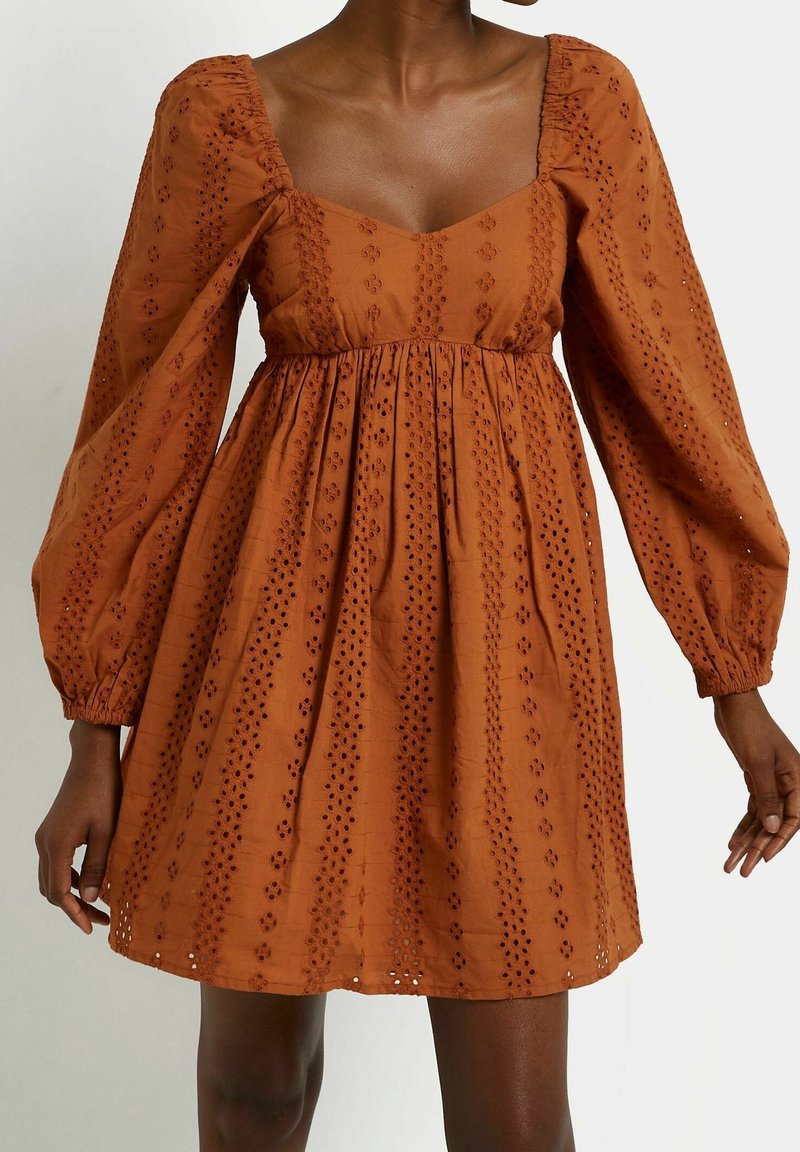 Robe longue à manches longues marron avec broderie ajourée, décolleté carré, taille froncée et jupe évasée portée par une personne debout.
