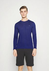 Tengerészkék hosszú ujjú ing puha anyagból, kerek nyakkivágással és finom logóval a mellkasnál, fekete rövidnadrággal párosítva.