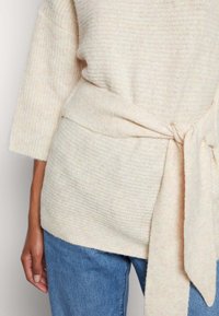 Pull beige en maille texturée avec manches trois-quarts et ceinture nouée à la taille, associé à un jean en denim bleu.