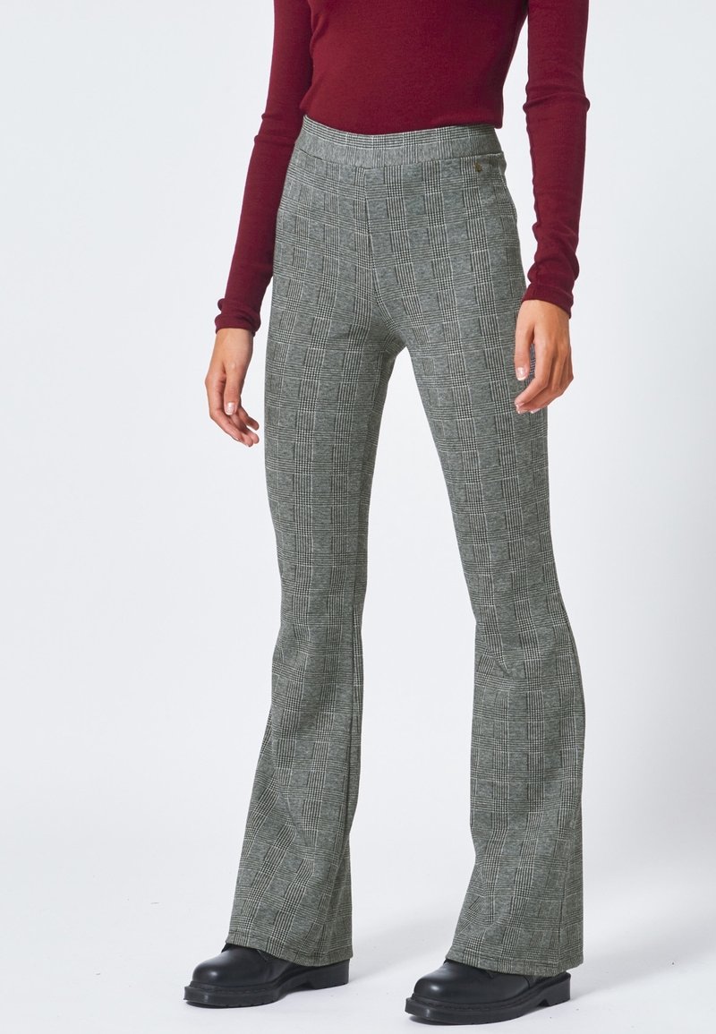 Femme portant un pantalon évasé à carreaux gris, des chaussures noires et un haut bordeaux cintré à manches longues, debout devant un fond clair uni.